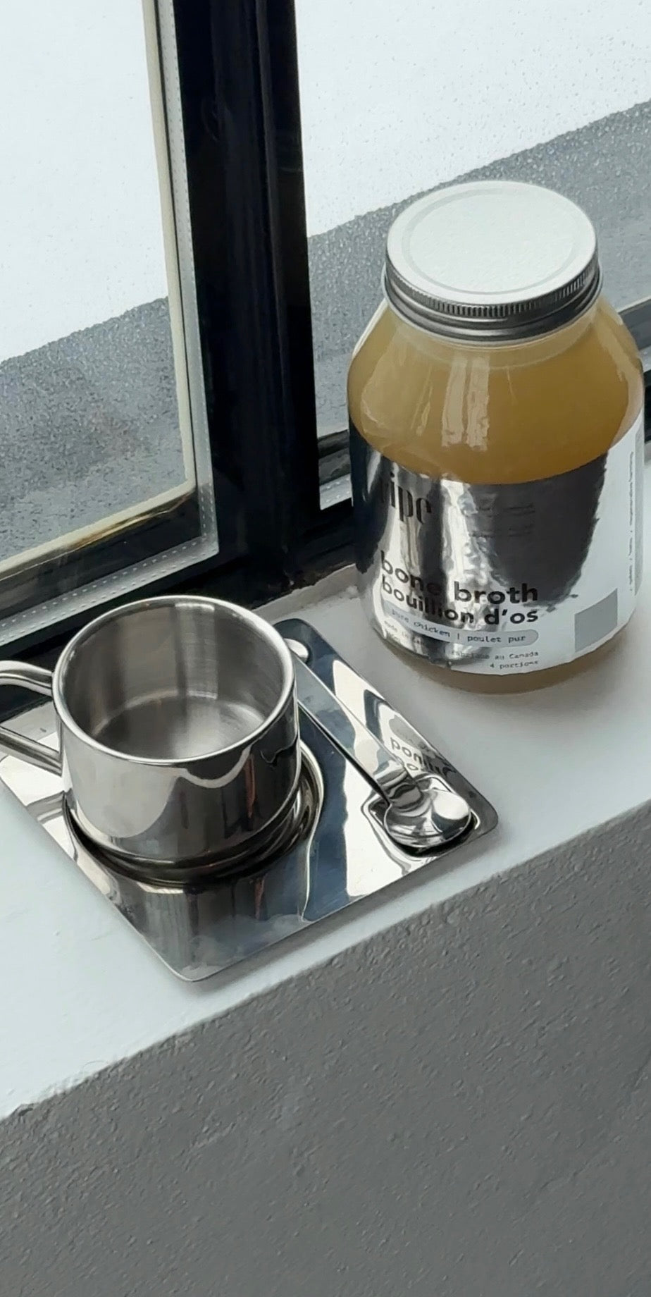 Jar of broth and metal cup on a reflective surface with a window in the background